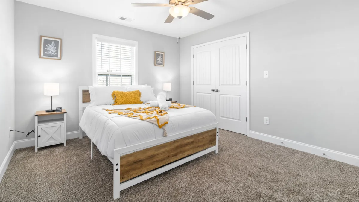 A sunlit bedroom with a comfortable bed adorned with a cheerful yellow throw and decorative pillows. A nightstand with a glowing lamp and art on the walls complete the cozy, inviting ambiance, suggesting restful mornings and peaceful evenings.