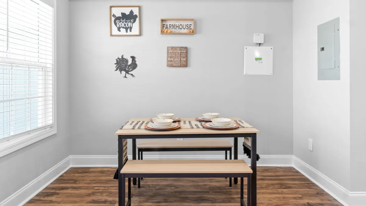 A welcoming dining table is placed near a window, with wall decorations and soft lighting fostering a warm, social atmosphere. The window offers a glimpse of natural light, hinting at the connection between indoor and outdoor spaces.