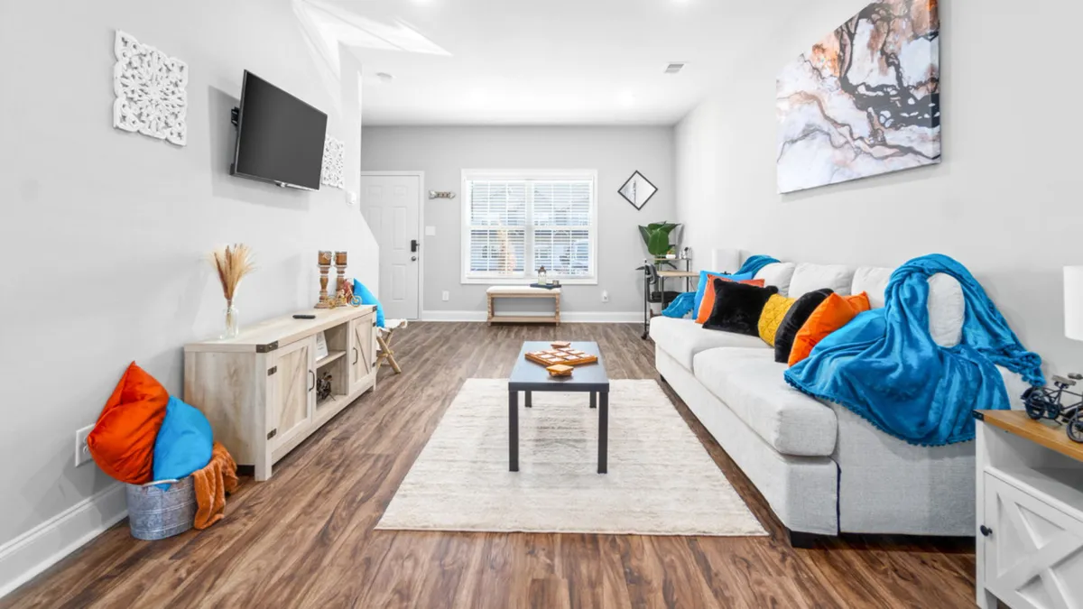 This image shows a cozy living room with a sofa adorned with colorful blankets, a coffee table, wall art, and a television. The space feels inviting and is set up for relaxing or entertaining guests, with seating and decor that promote a sociable atmosphere.