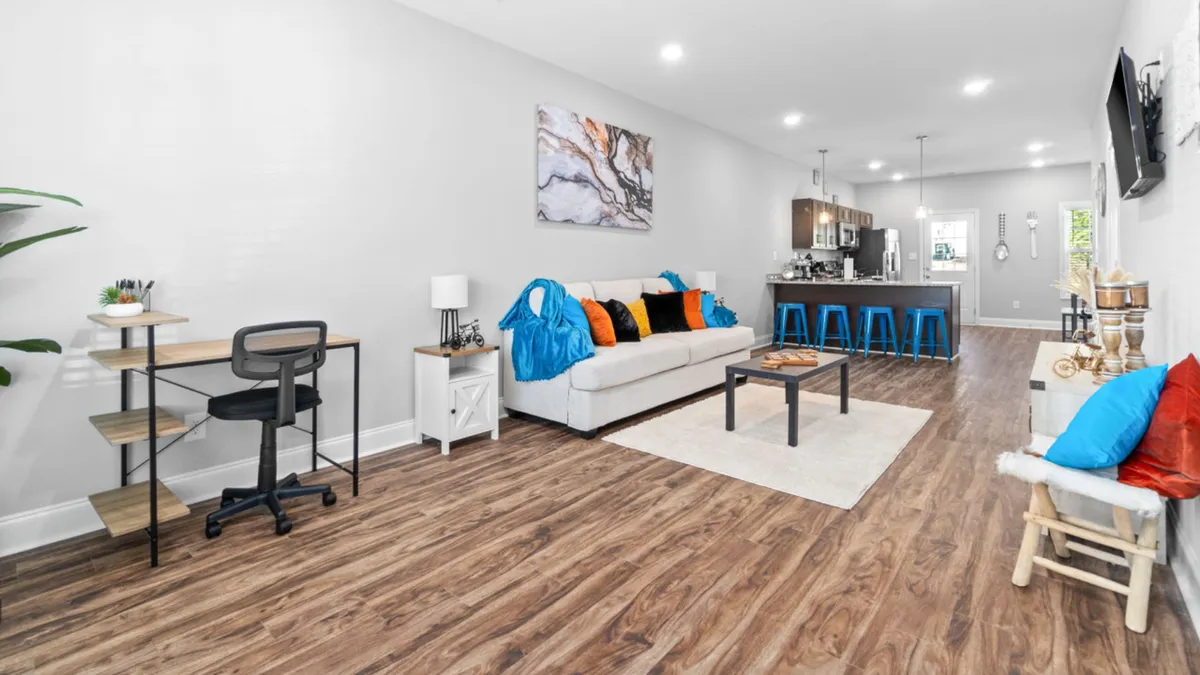 The image showcases a sunlit, stylish living area with a plush sofa adorned with colorful cushions, eye-catching artwork displayed on the wall, and a modern dining area in the background. The scene evokes a sense of comfort and warmth, emphasizing both social and relaxing spaces ideal for gatherings or downtime.