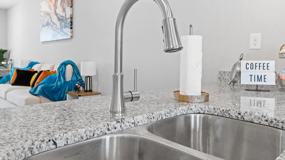 A cheerful kitchen scene featuring a modern sink, granite countertop, and a 'coffee time' sign, with a glimpse into a bright living area. The space suggests a welcoming atmosphere for morning routines or enjoying a break.