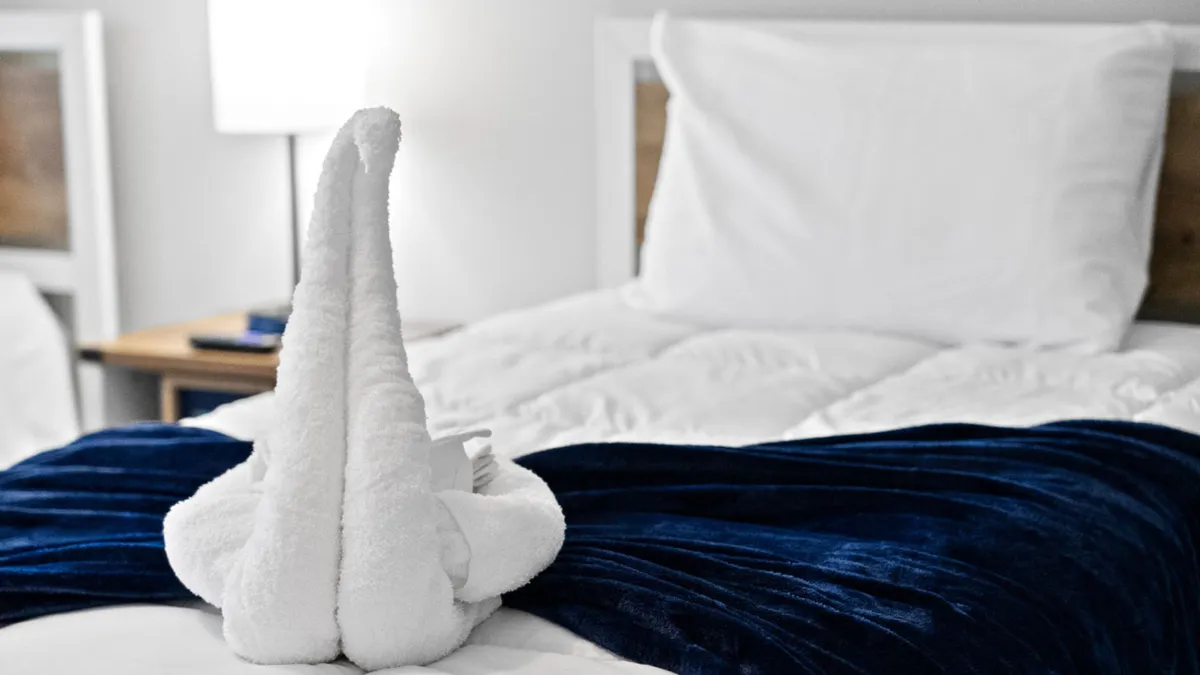 This image features a beautifully made bed with crisp white bedding, a plush blue velvet throw, and a towel elegantly folded into the shape of a swan. Soft lighting from a bedside lamp enhances the serene atmosphere, and the arrangement suggests a special effort made for a romantic evening.