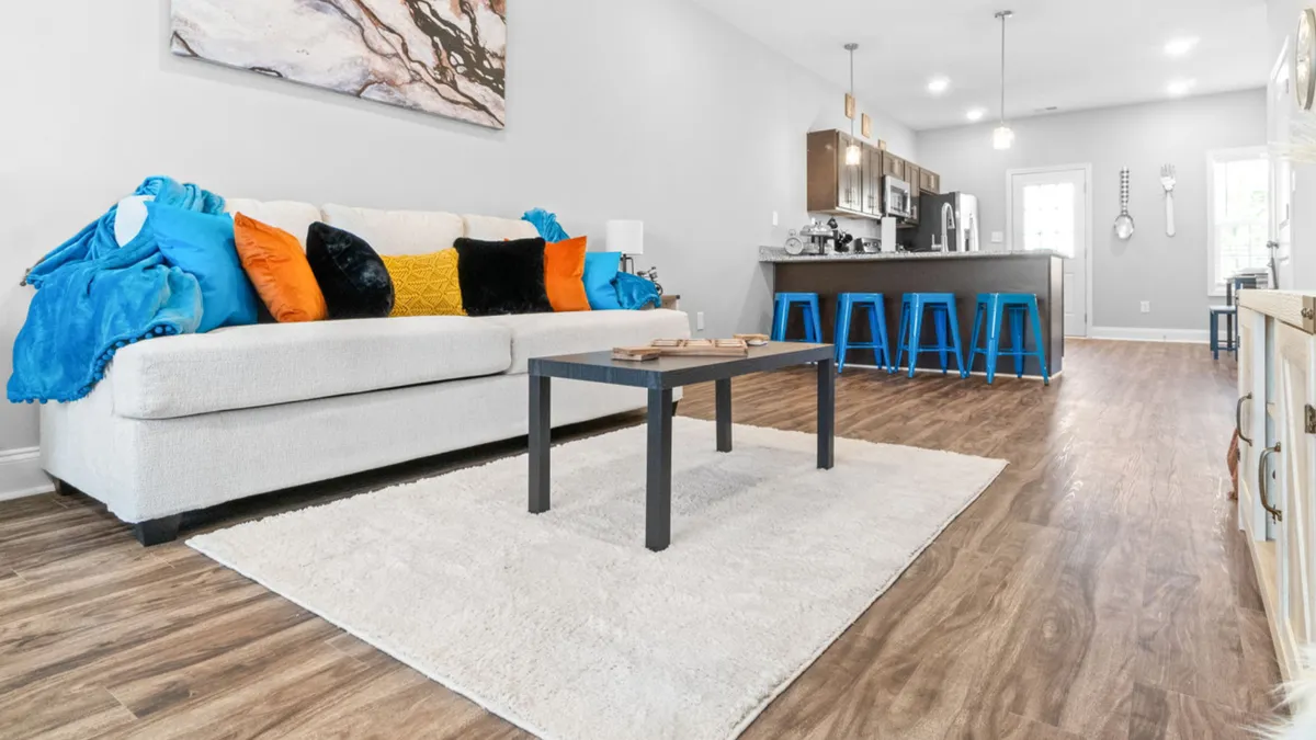 A spacious, inviting living room featuring a plush sofa adorned with colorful cushions, a central coffee table, and a view into the kitchen area with bar stools. The open layout encourages socializing and relaxation, while warm lighting and stylish decor enhance the welcoming atmosphere.